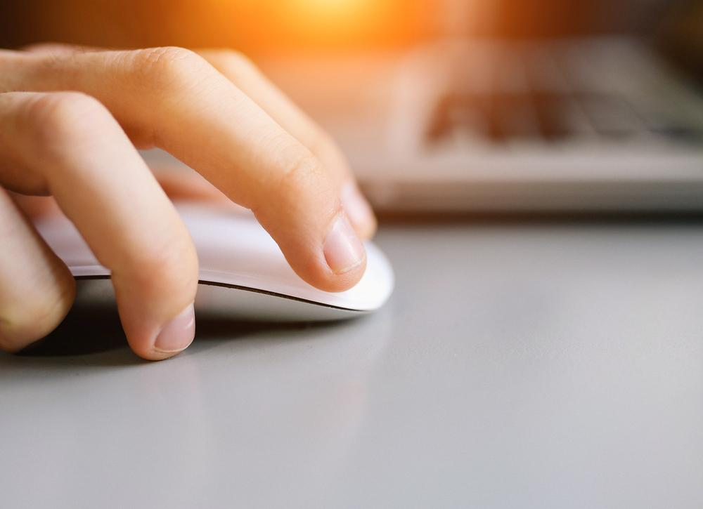 A close-up of a persons hand using a white computer mouse next to a laptop, with sunlight shining in the background.